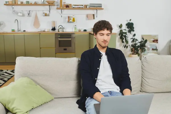 Curly-haired man sits on a sofa, attentively focused on his laptop in contemporary living space. — Stock Photo