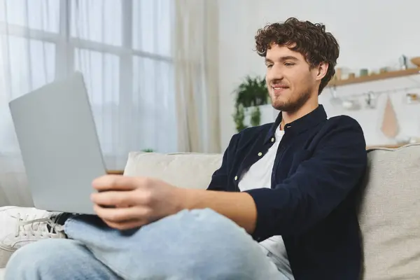 Jovem bonito com cabelo encaracolado relaxa no sofá, engajando-se com seu laptop em um espaço acolhedor. — Fotografia de Stock