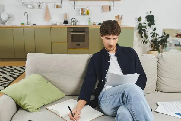 A young man with curly hair sits comfortably in his stylish home, focusing on his notes and writing. — Stock Photo
