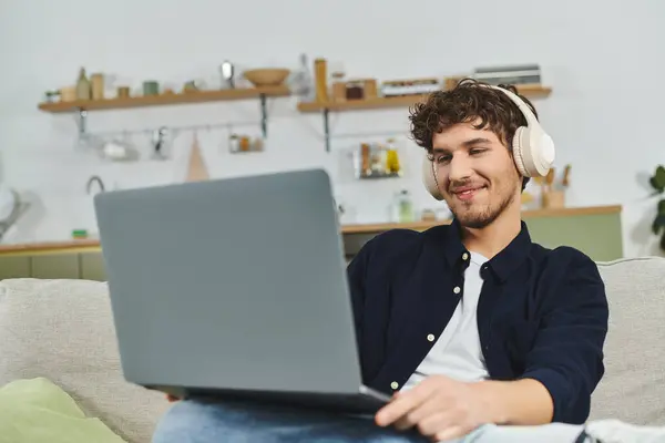 Bonito homem encaracolado relaxa no sofá com fones de ouvido, focado em seu laptop em espaço de vida elegante. — Fotografia de Stock