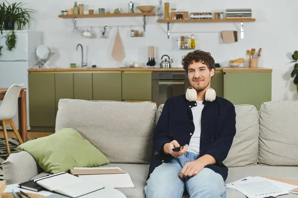 El hombre de pelo rizado se relaja en el espacio de vida moderno, sosteniendo un remoto y sonriente contentamiento. - foto de stock