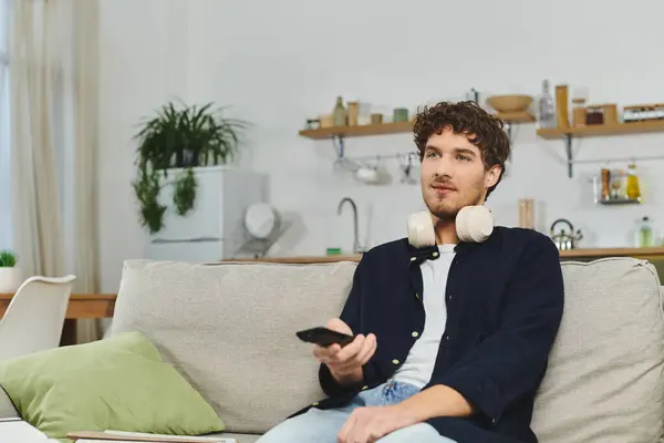 Curly-haired young man sits comfortably at home with headphones on, holding a remote control. — Stock Photo