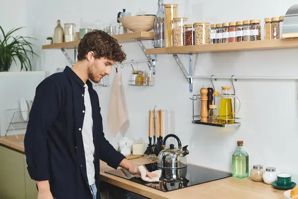 Jovem bonito com cabelo encaracolado limpa seu fogão em uma cozinha elegante cheia de luz. — Fotografia de Stock