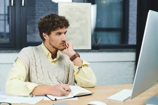 Curly-haired young man in a sweater vest focuses on a computer while taking notes at work. — Stock Photo