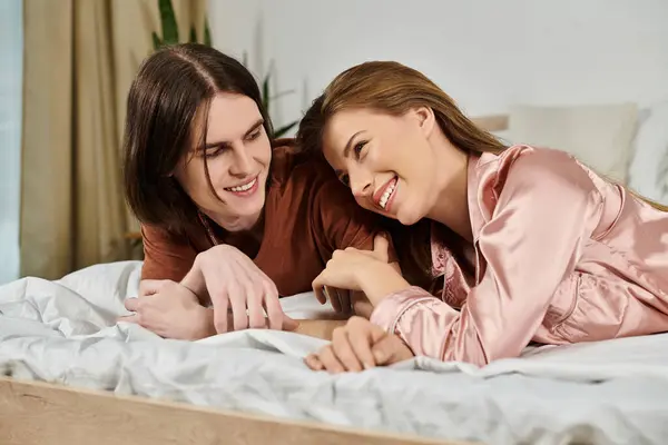 Two young individuals lie on a bed, smiling and engaging with each other in a cozy interior setting. — Stock Photo
