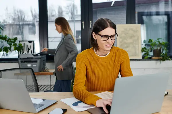 A young person in an orange sweater works on a laptop while a colleague organizes documents. — Stock Photo