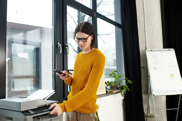 A young person with a smartphone near a printer in a chic office, focused on tasks. — Stock Photo