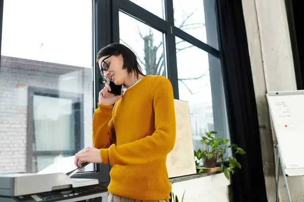 A young individual in a chic sweater talks on the phone near a large window while handling papers. — Stock Photo
