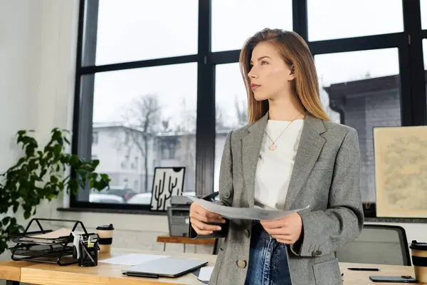 In a stylish office, a young chic individual stands thoughtfully holding documents while gazing out. — Stock Photo