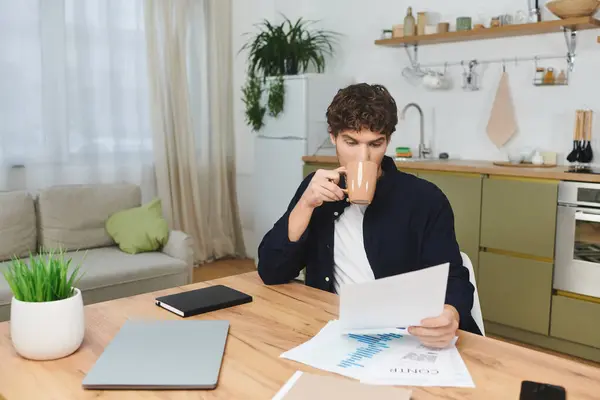 Handsome young man with curly hair sips coffee and studies papers in a modern home setting. — Stock Photo