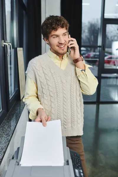 Um jovem homem encaracolado em um colete de camisola conversa ao telefone enquanto trabalhava em um ambiente de escritório. — Fotografia de Stock