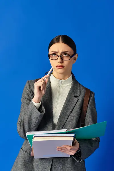 Una giovane donna in occhiali sembra premurosa, tenendo quaderni e una penna su uno sfondo blu. — Foto stock