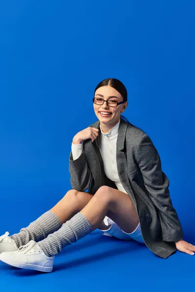 Happy young woman in a chic outfit sits on the floor against a blue backdrop. — Stock Photo