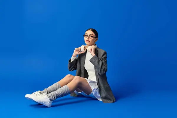 A young woman in a playful outfit sits on the floor, expressing emotions against a blue backdrop. — Stock Photo