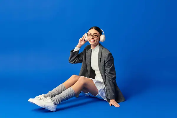 A young woman in a stylish outfit sits on the floor smiling while listening to music. — Stock Photo
