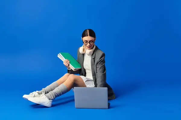 Eine junge Frau mit freudigem Gesichtsausdruck ist mit ihrem Laptop beschäftigt, während sie Bücher in der Hand hält. — Stockfoto