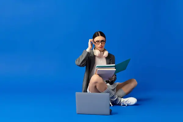 Jeune femme assise sur fond bleu, tenant des livres et regardant réfléchi tout en étudiant. — Photo de stock