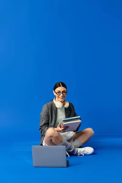 Emotional young woman sits cross legged with books and a laptop on a bright blue floor. — Stock Photo