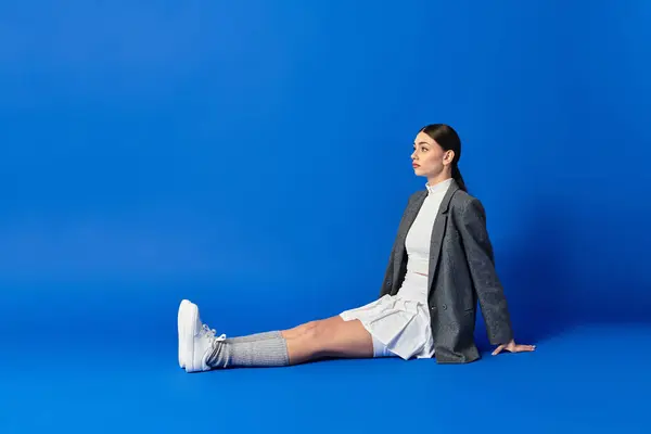 A young woman in a chic outfit sits on the ground, deep in thought against a blue backdrop. — Stock Photo
