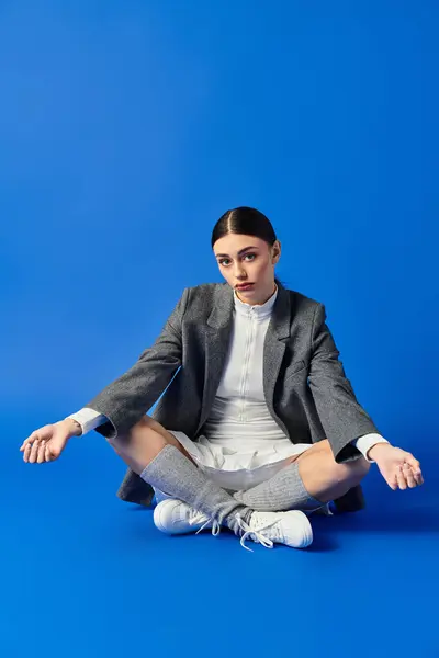 A young woman in a chic outfit sits calmly against a bold blue backdrop, exuding emotion. — Stock Photo