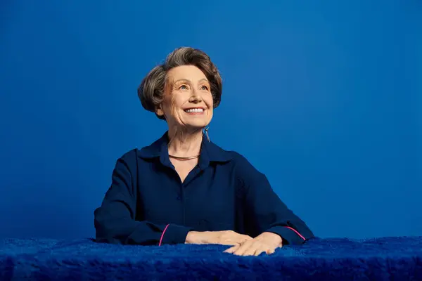 A beautiful woman in her 60s poses confidently in trendy attire against a striking blue background. — Stock Photo