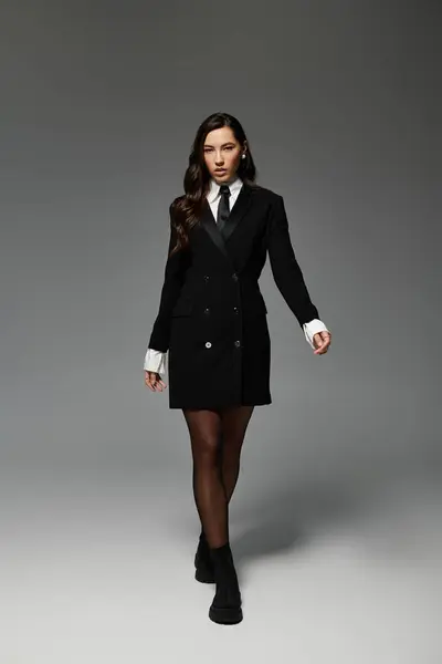 A beautiful young woman confidently poses in a sleek outfit against a grey backdrop, exuding charm. — Stock Photo