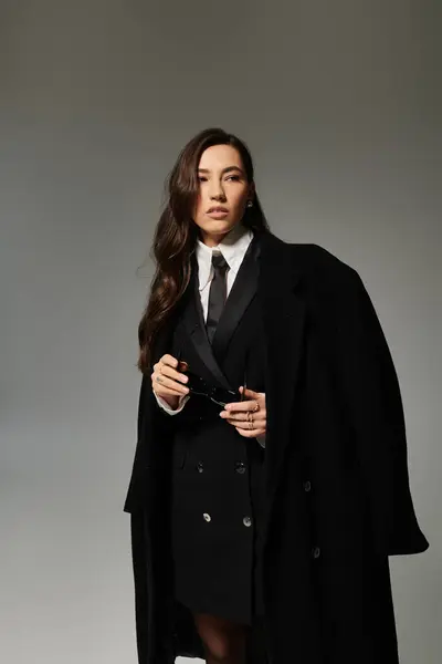 Una hermosa joven con el pelo castaño largo posa elegantemente en ropa elegante contra el gris. - foto de stock