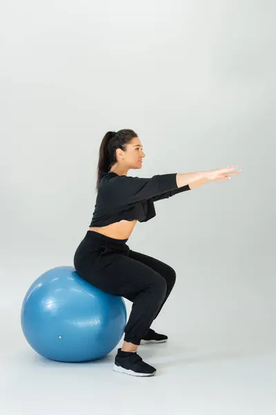 Mujer en cuclillas en una bola de estabilidad en un espacio interior brillante, mostrando fuerza y enfoque. - foto de stock