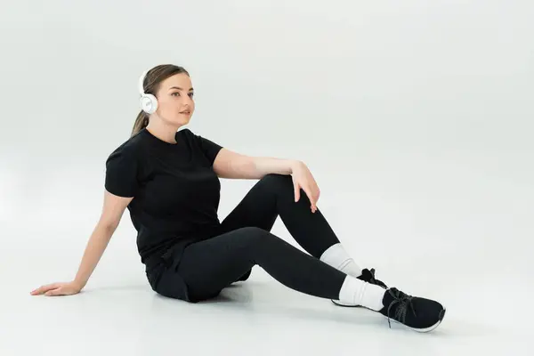 A strong and beautiful woman sits on the floor, wearing sportswear and headphones, taking a break. — Stock Photo