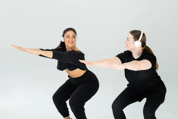 Two strong women perform synchronized squats while wearing headphones in a vibrant fitness studio — Stock Photo