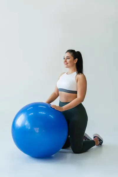 Mujer en forma se centra en su entrenamiento mientras se arrodilla junto a una pelota de fitness azul en un gimnasio brillante. - foto de stock