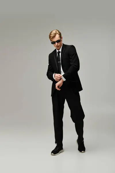 A young man with stylish features elegantly adjusts his sleeves while wearing a sleek black suit. — Stock Photo