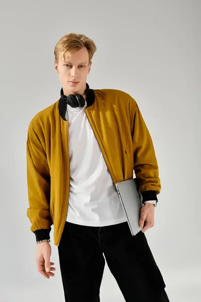 A young handsome man stands confidently holding a laptop while dressed in a trendy jacket. — Stock Photo