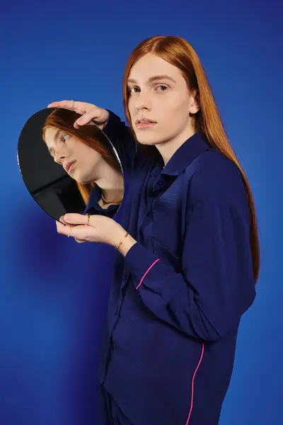 A fashionable young man with long red hair admires his reflection in a sleek round mirror. — Stock Photo