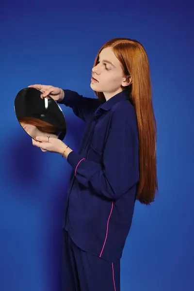 A young queer man with long red hair poses with a mirror against a vibrant blue backdrop. — Stock Photo