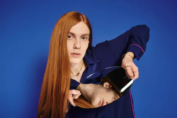 A young man with long red hair thoughtfully examines his unique style in a mirror. — Stock Photo