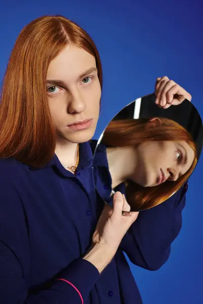 A young man with long red hair gazes thoughtfully into a mirror, contemplating self expression. — Stock Photo