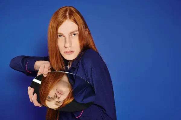 Un giovane uomo queer con lunghi capelli rossi colpisce una posa audace con uno specchio, mostrando il suo stile. — Foto stock