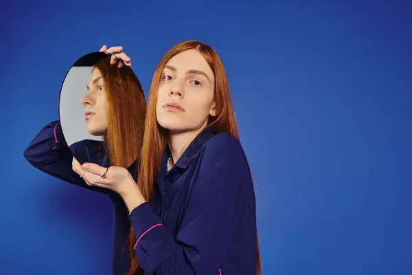 Un giovane uomo con lunghi capelli rossi posa fiducioso con uno specchio alla moda che lo riflette. — Foto stock