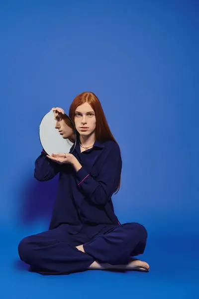 Un giovane alla moda con lunghi capelli rossi contempla il suo riflesso in un ambiente elegante. — Foto stock