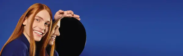 A young queer man beams with joy while checking his reflection against a circular mirror. — Stock Photo