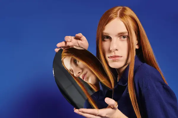 A young queer man with long red hair poses with a mirror against a bold blue background. — Stock Photo