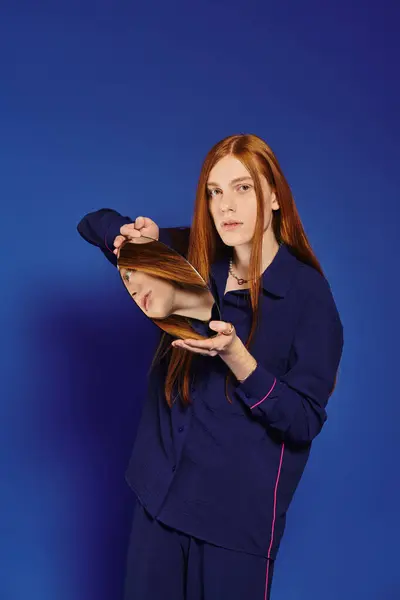 A young queer man with long red hair holds a mirror, his face surrounded by deep blue. — Stock Photo