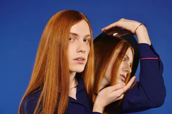 A stylish young man admires his long red hair in a mirror with a bright blue backdrop. — Stock Photo