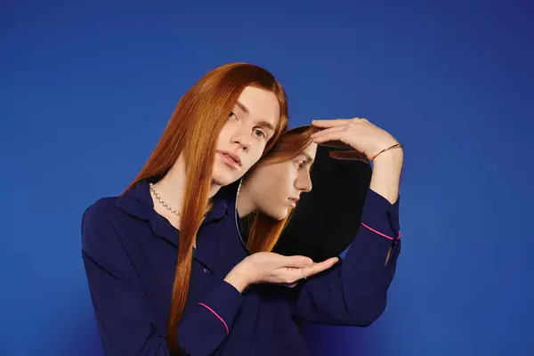 Un giovane uomo queer con lunghi capelli rossi guarda pensieroso in uno specchio, meditando la sua identità. — Foto stock