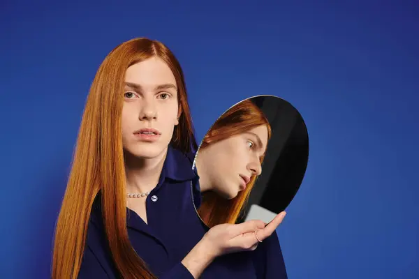 Un giovane uomo con lunghi capelli rossi guarda in uno specchio tenuto in mano su uno sfondo blu vibrante. — Foto stock