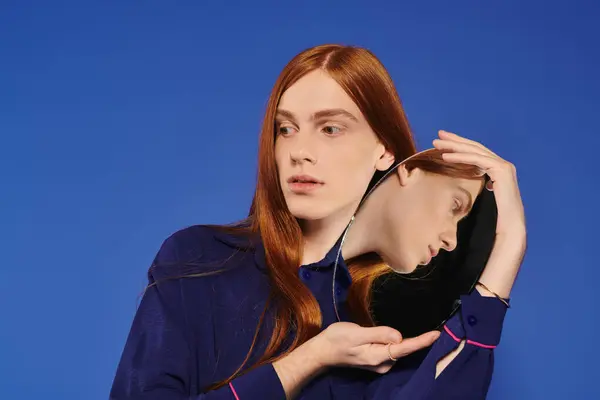 Un bel giovane uomo con lunghi capelli rossi ammira il suo riflesso in uno specchio blu unico. — Foto stock