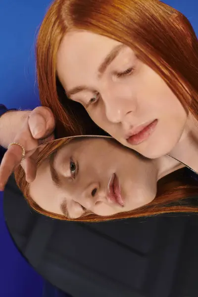 Un giovane uomo queer con lunghi capelli rossi si impegna pensierosamente con uno specchio, esplorando l'identità di sé. — Foto stock