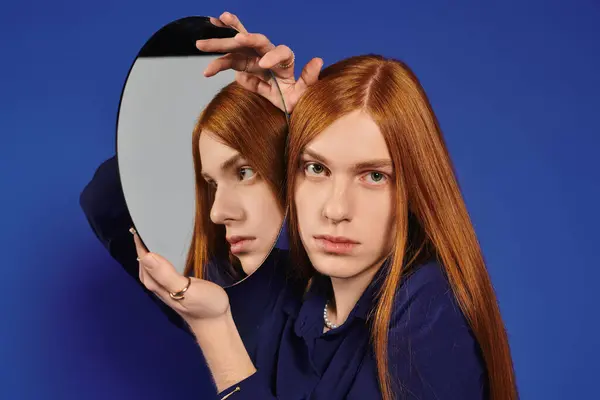 A young queer man with long red hair stares at his reflection in a blue backdrop. — Stock Photo