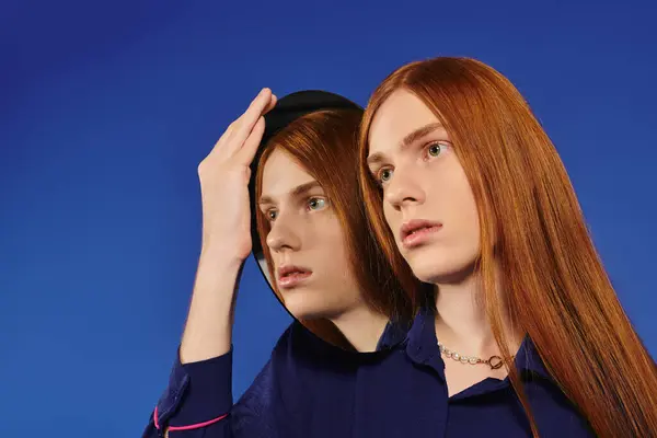 A young queer man with long red hair poses stylishly against a blue backdrop. — Stock Photo